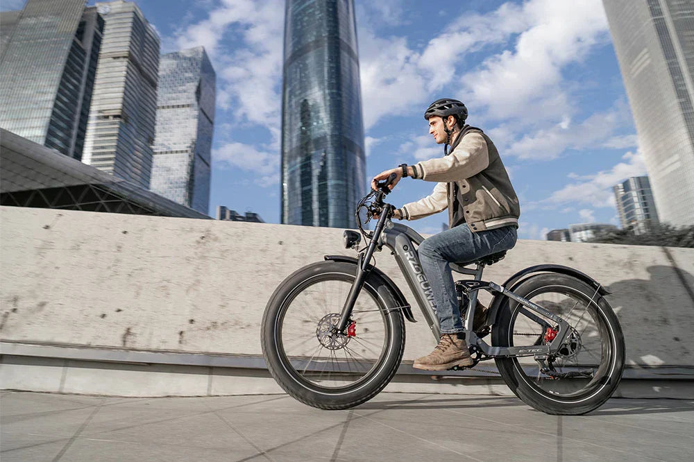 Low-angle downtown city lifestyle shot of a helmeted man riding gunmetal grey RZOGUWEX S7 fat tire e-bike on urban pavement with modern skyscrapers in background.