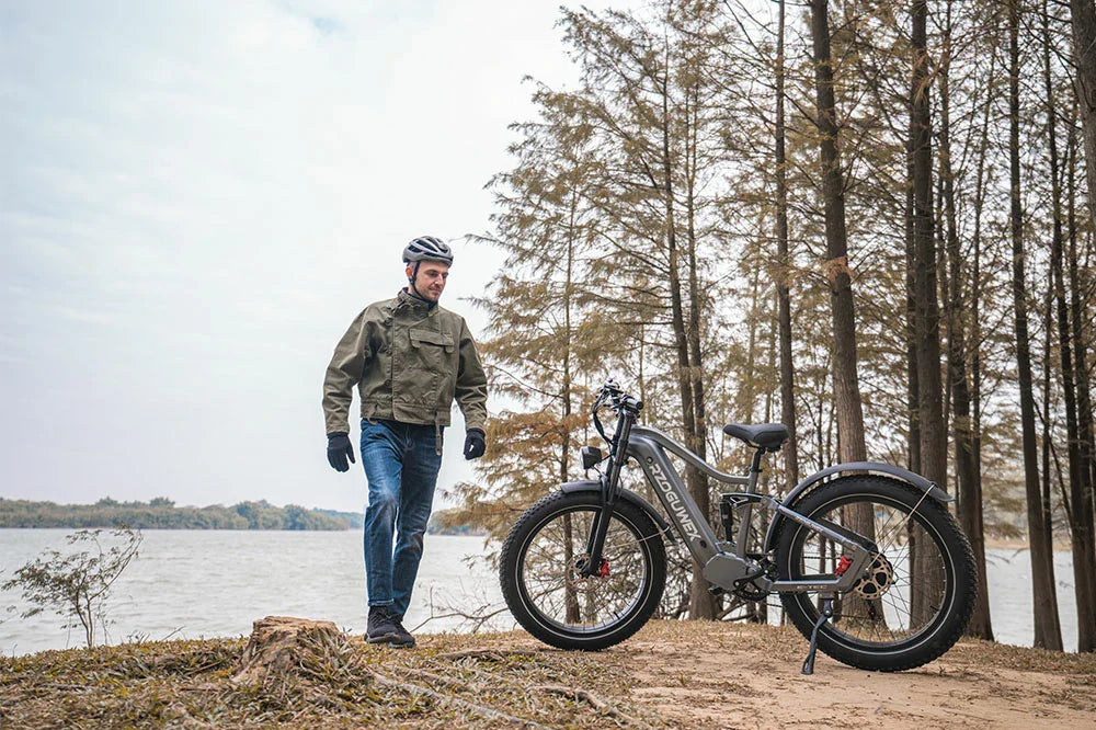 Calm waterside lifestyle shot of helmeted man walking beside matte grey RZOGUWEX S7 fat tire electric bike on lakeside woodland shore.