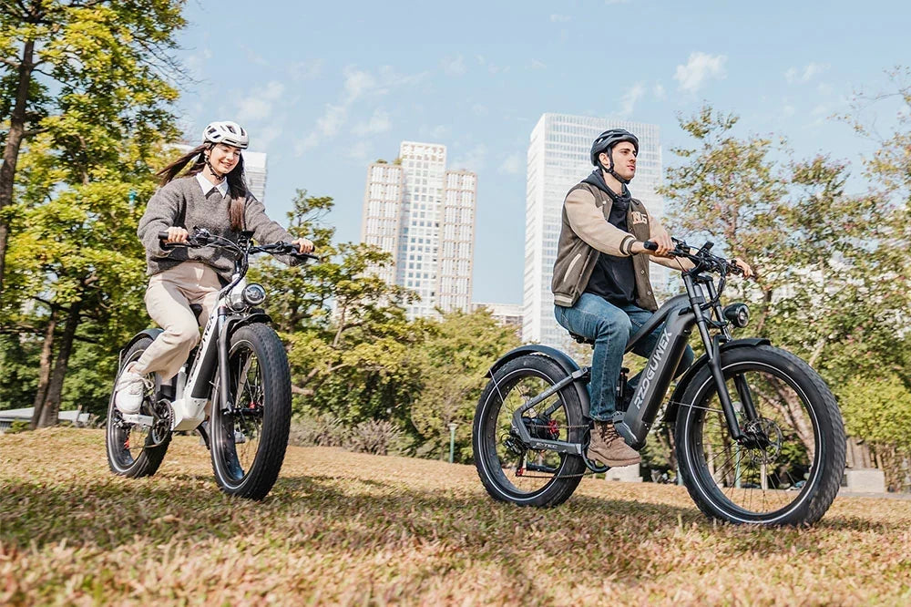 Urban park lifestyle shot of two helmeted riders cruising white and gunmetal grey RZOGUWEX S7 fat tire e-bikes across sunlit grass with modern glass skyscrapers and lush green trees in the background.