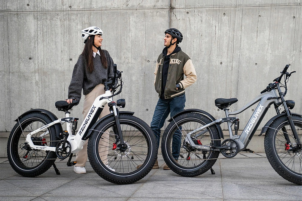 Urban lifestyle photo, young couple standing beside white and grey RZOGUWEX S7 full suspension fat tire electric bikes against concrete wall.