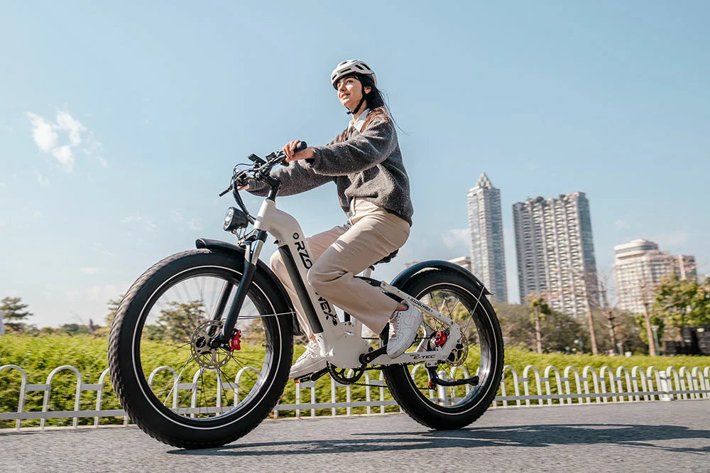 This is the RZOGUWEX S9, a white fat-tire electric bike, being ridden by a woman wearing a helmet in an urban park setting with modern high-rise buildings in the background.