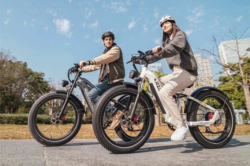 A man and woman ride dark grey and white RZOGUWEX S7 fat tire e-bikes side by side in a sunny urban park setting.