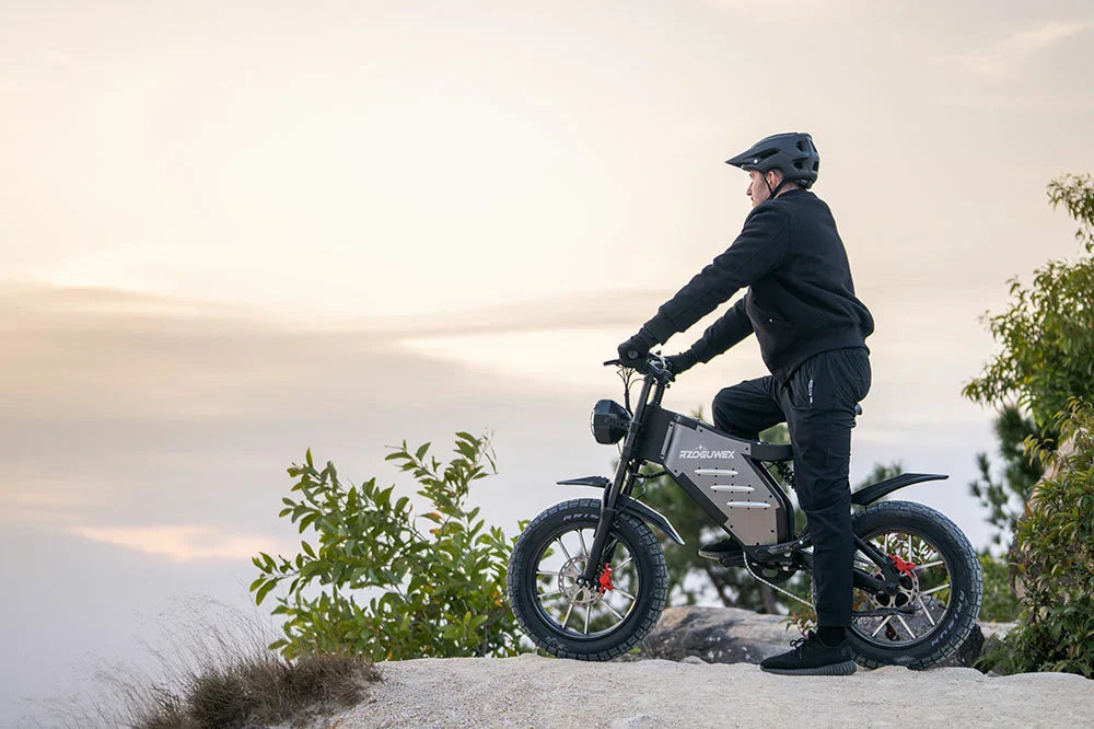Soft sunset cliffside lifestyle shot of helmeted male rider perched on matte grey RZOGUWEX X5S retro fat tire e-bike overlooking hazy pastel sky atop rocky coastal bluff.