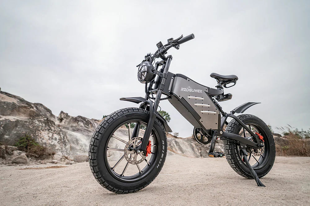 Front three-quarter static shot of matte grey RZOGUWEX X5S retro fat tire electric dirt bike parked on rocky desert terrain under overcast sky.