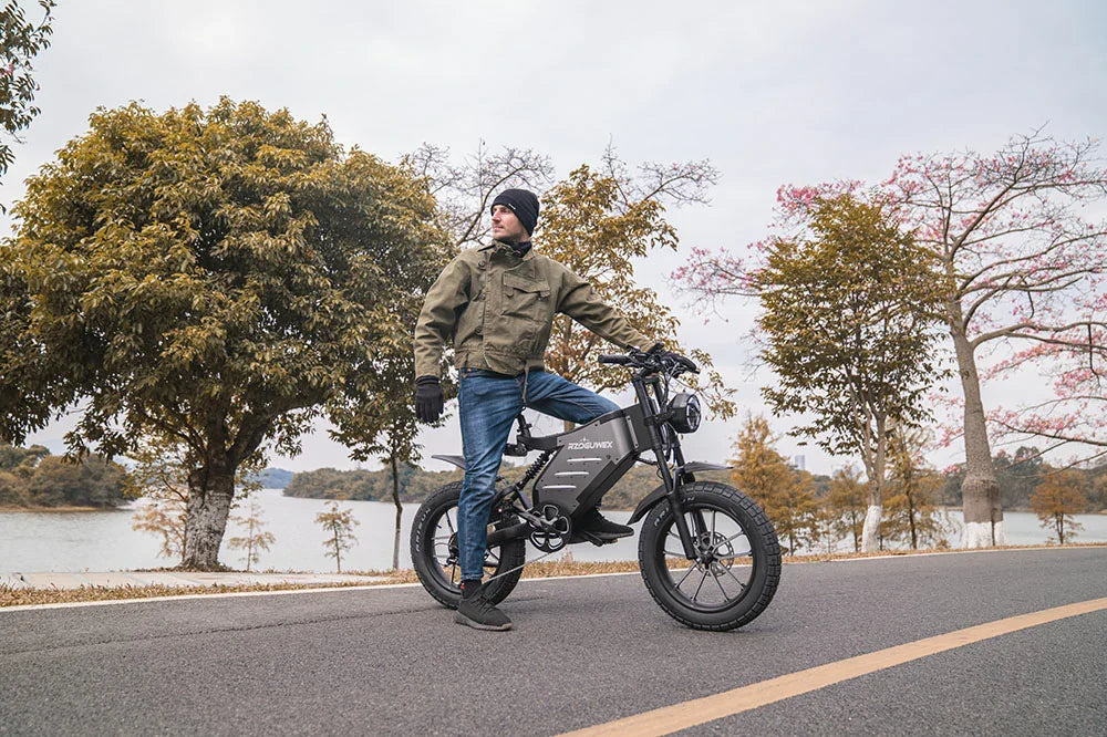Lakeside roadside lifestyle shot of a man posing beside matte grey retro RZOGUWEX X5S fat tire electric cruiser bike, lined with autumn foliage trees and calm lake waters.